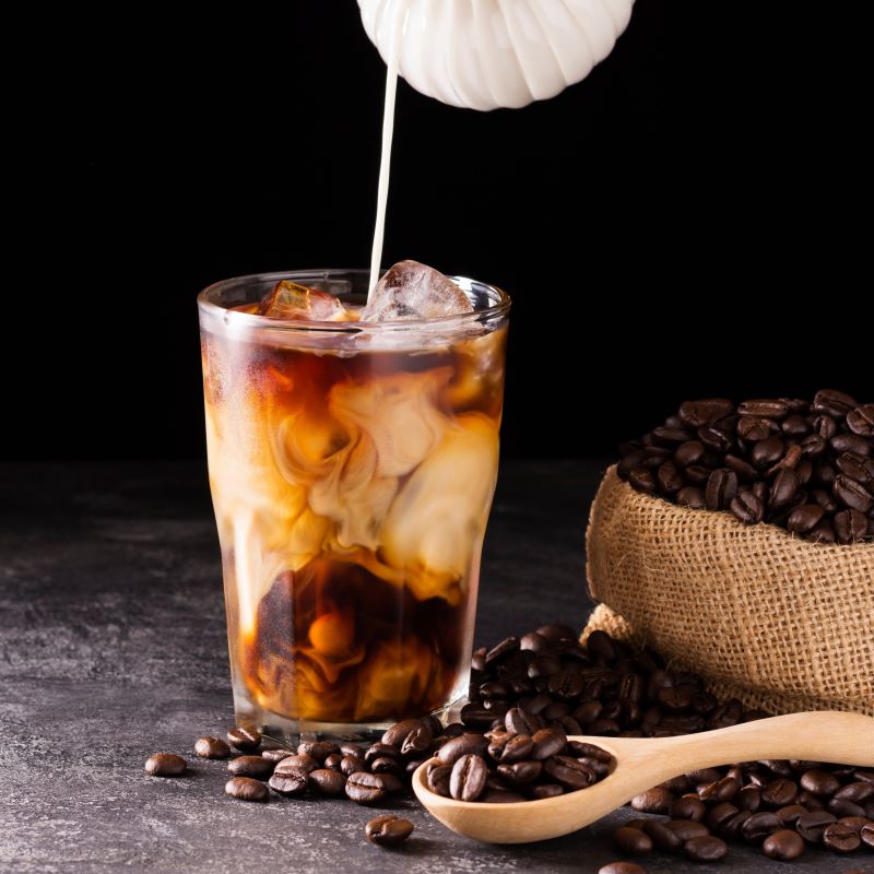Iced coffee being poured into a glass with coffee beans around on a dark surface
