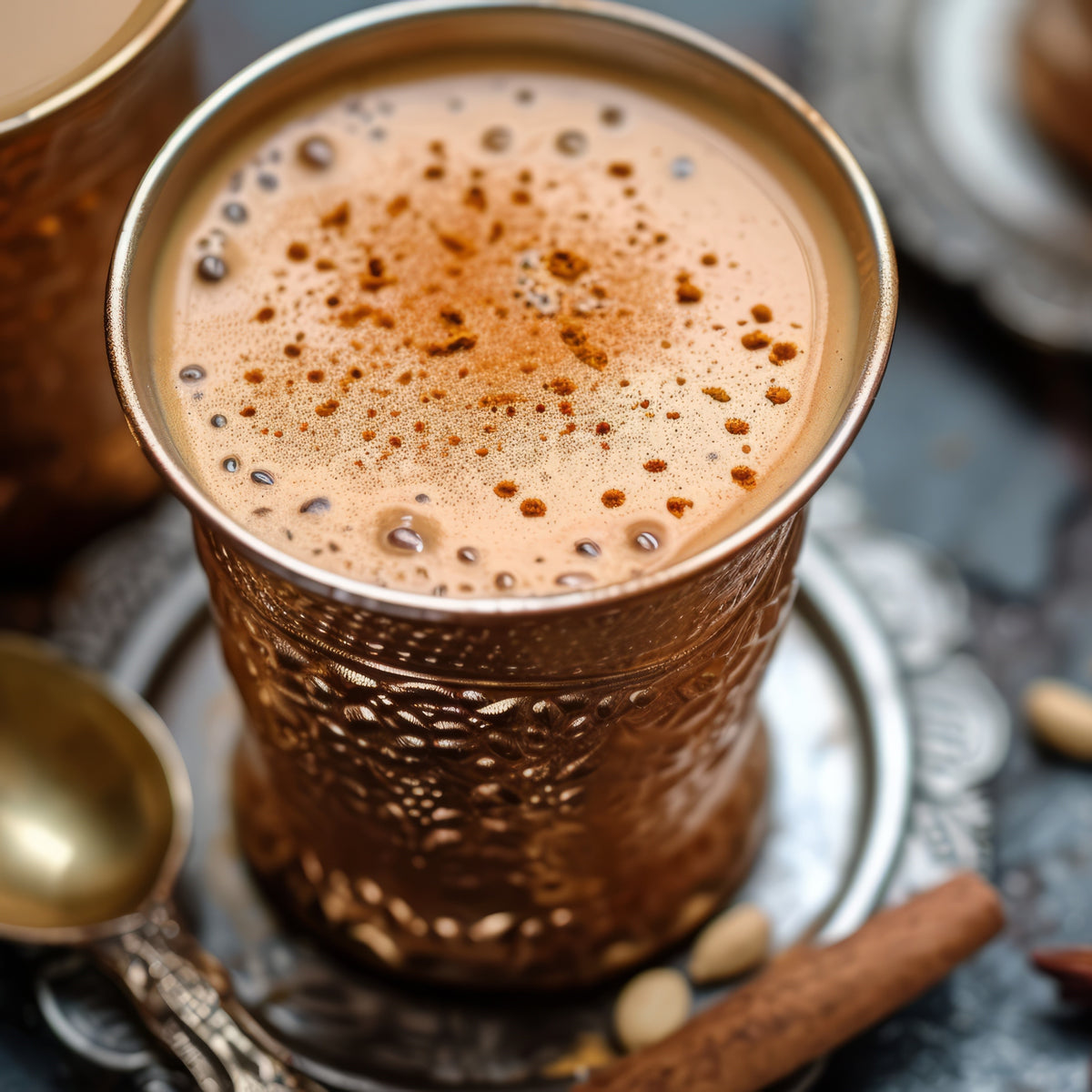 Copper cup filled with a frothy beverage on a dark surface with cinnamon sticks and a spoon.