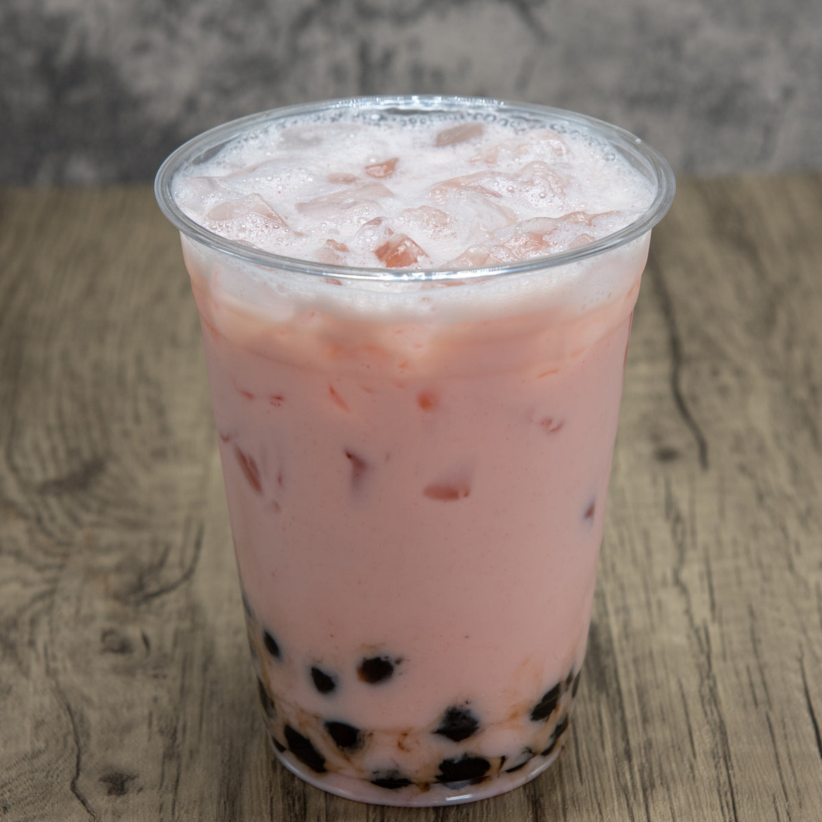 Pink bubble tea with black tapioca pearls in a clear plastic cup on a wooden surface.